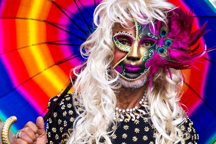 A man wearing a wig and a mask poses for a photograph while holding a colourful parasol