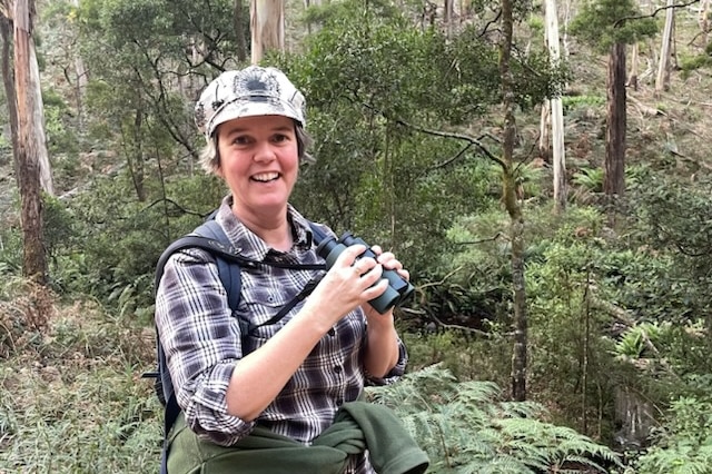 A woman smiles at the camera while holding binoculars, she is in a forest