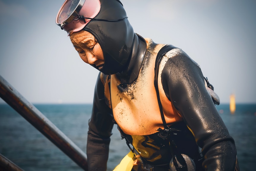 A woman wearing scuba gear, with an orange vest and goggles on her head look down as she gets into the water.