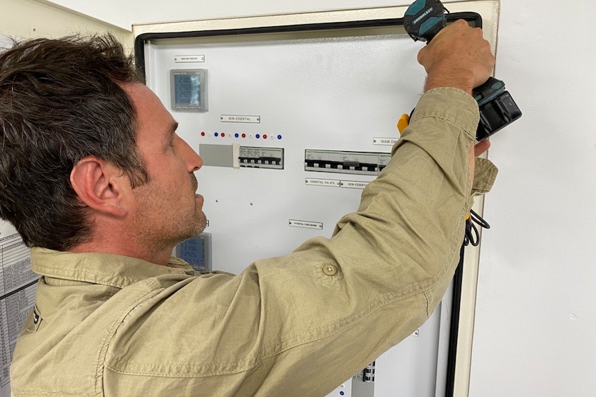 A man with short dark hair, wearing a long-sleeved work shirt, uses a drill on an electrical switchboard.