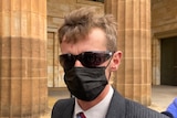 A man with short brown hair wearing a black face mask, sunglasses and a suit