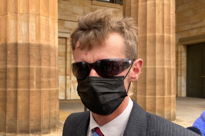 A man with short brown hair wearing a black face mask, sunglasses and a suit