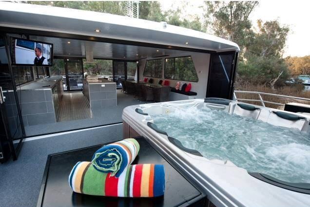 The upper deck of a houseboat with a largeoutside  spa pool and a covered modern kitchen and dining space with a large TV