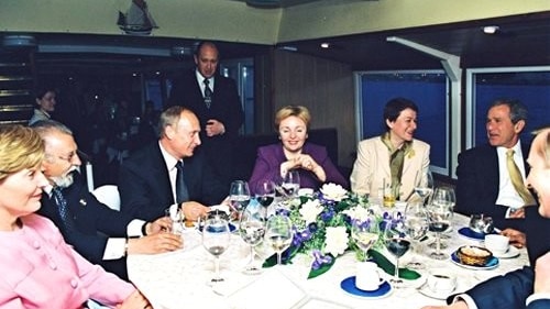 Vladimir Putin, George Bush and other guests sit at a round dinner table inside a boat. A man in a suit watches on