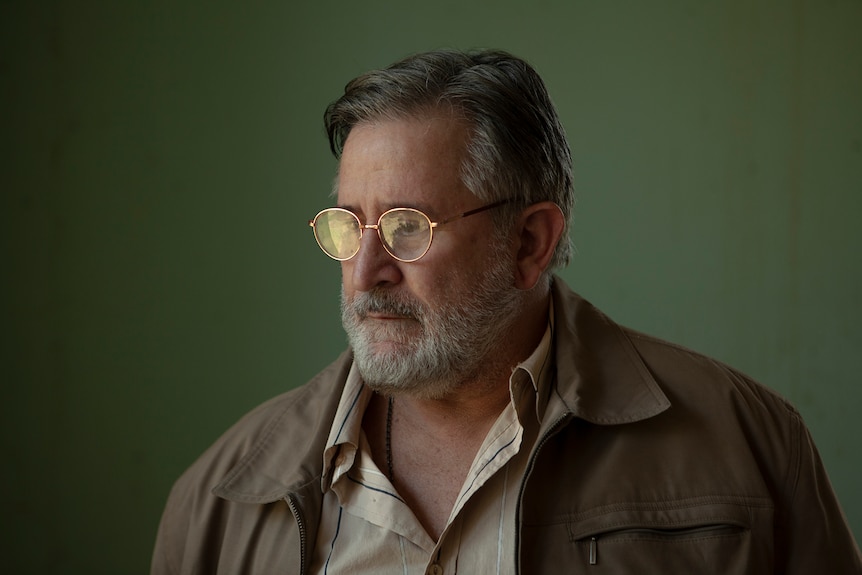 Middle-aged man with a grey beard and wearing glasses and a tan jacket looks downcast in front of an olive green wall.
