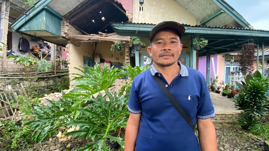 A man wearing a hat standing in front of a destroyed house due to an earthquake