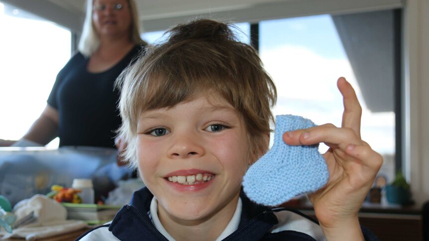 A young boy holds a knitted baby bootie up to the camera.