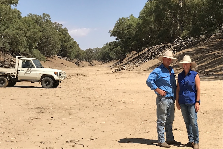 two people standing in red sand on a dry creek bed with a white car inn the background and a blue sky.