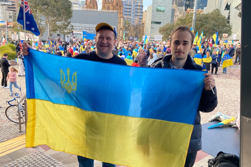 Two men holding a Ukrainian flag. One is wearing a cap and the other has his hair tied back.