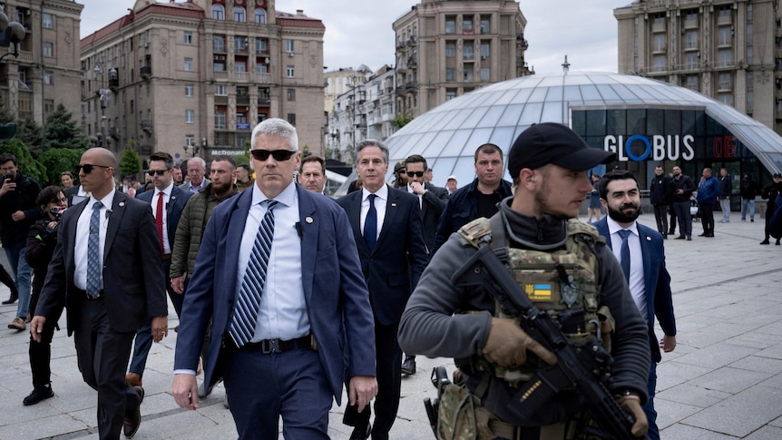 An older white man in a suit looks serious as he walks through a town square surrounded by security.