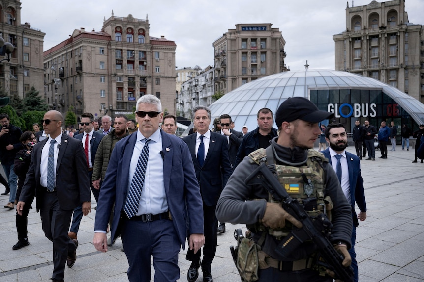 An older white man in a suit looks serious as he walks through a town square surrounded by security.