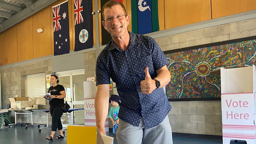 A man in a blue collared shirt and shorts places his voting paper in a ballot box, with a thumbs up gesture.