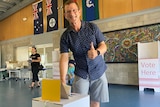 A man in a blue collared shirt and shorts places his voting paper in a ballot box, with a thumbs up gesture.