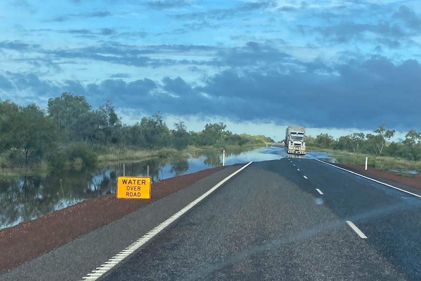 A truck driving on a remote highway with water on the road, with a yellow warning sign on one side of the road.