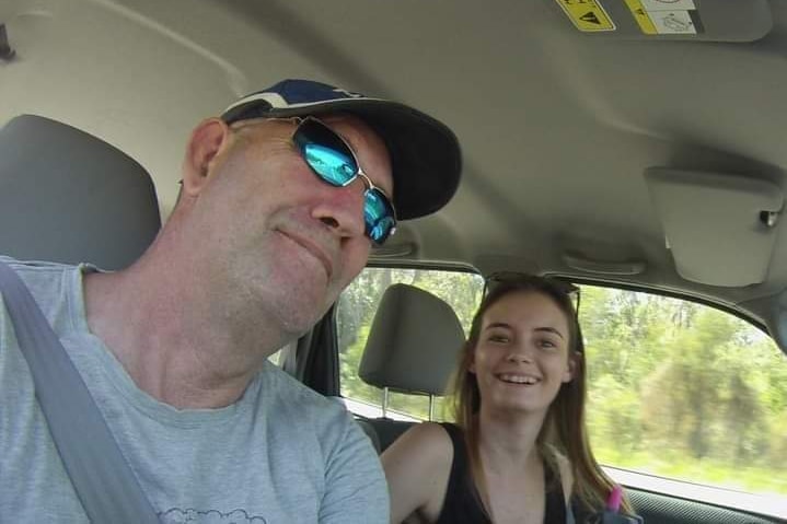 A man in a cap and reflective sunnies smiles in a car with a young woman beside him.