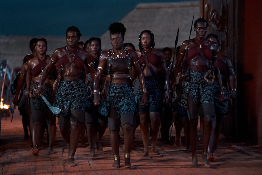 A group of Black soldiers marching.  They're wrapped blue skirts and shorts, red tops and plated armor holding swords and spears