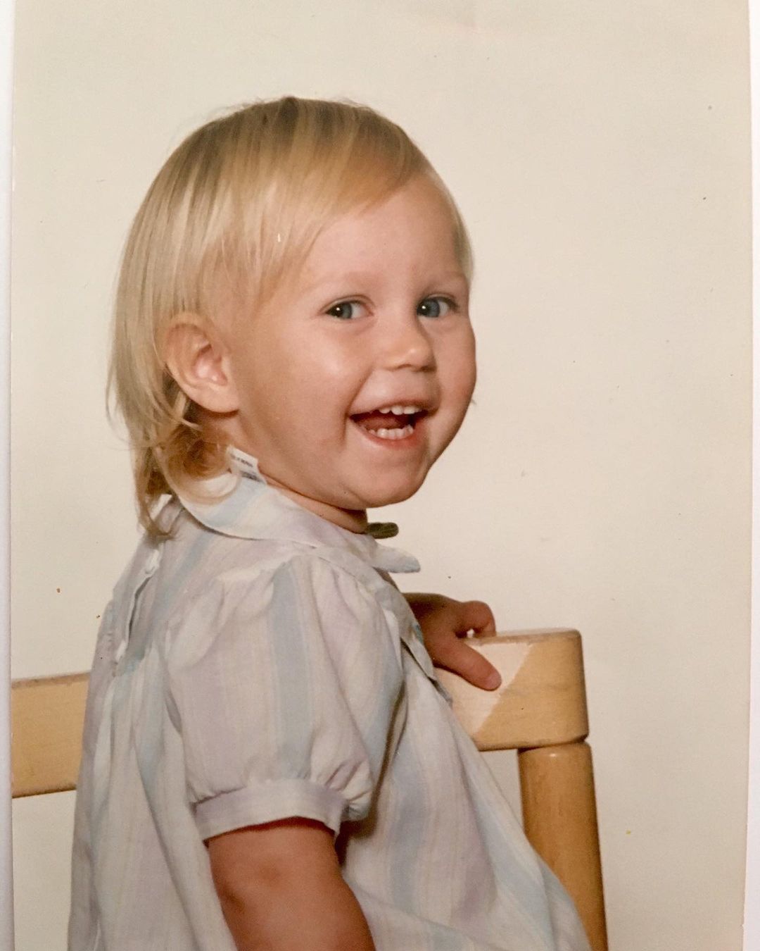A smiling blonde child sits sideways on a wooden chair, turning her head to the camera. She wears a pastel striped number