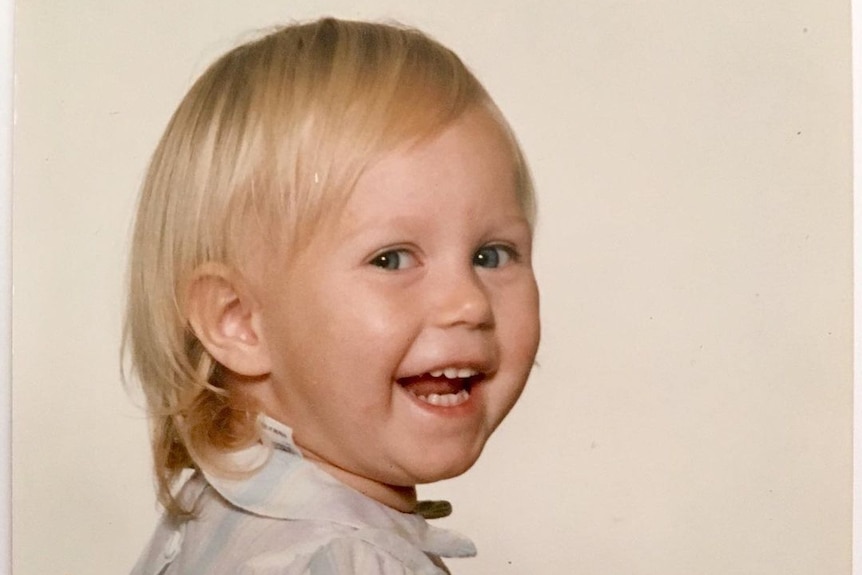 A smiling blonde child sits sideways on a wooden chair, turning her head to the camera. She wears a pastel striped number