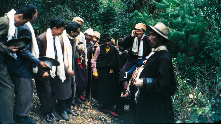 A group of men stand by as the Dalai Lama, in a scarf, glasses and hat, rides past