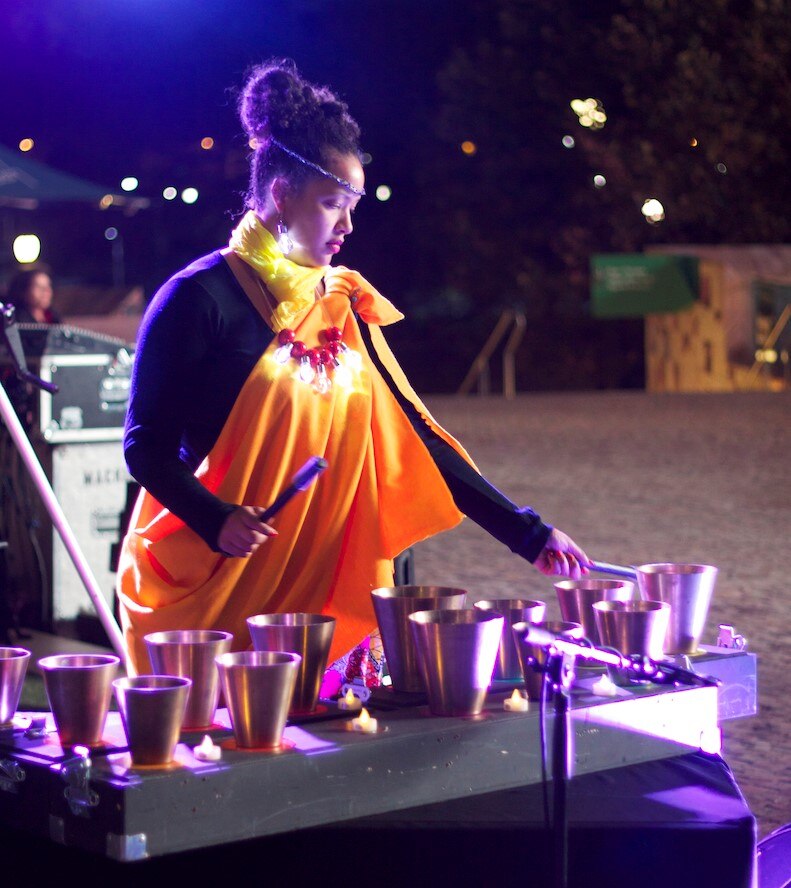 A woman in a bright yellow wrap dress stands outside leaning over a table with a series of copper containers she is playing