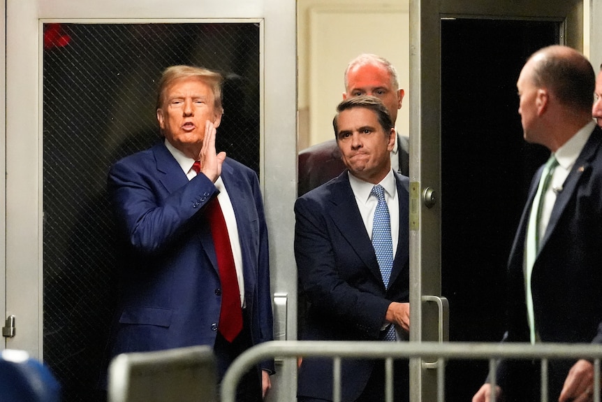 Donald Trump speaks while holding a hand next to his chin as he stands next to a doorway, which two men are walking through.