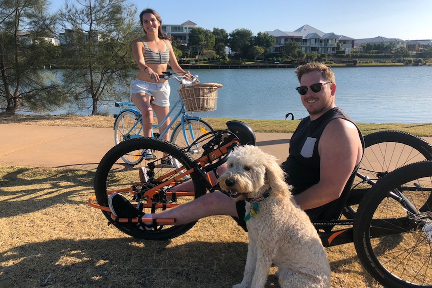 Paralympian Matt McShane smiles from his wheelchair - next to him are his dogs and behind him is his fiance Ella Sabljak