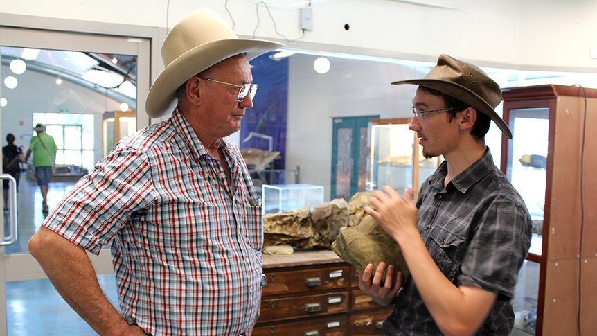 two men with bush hats on looking at each as the one on the right holds a large rock fossil