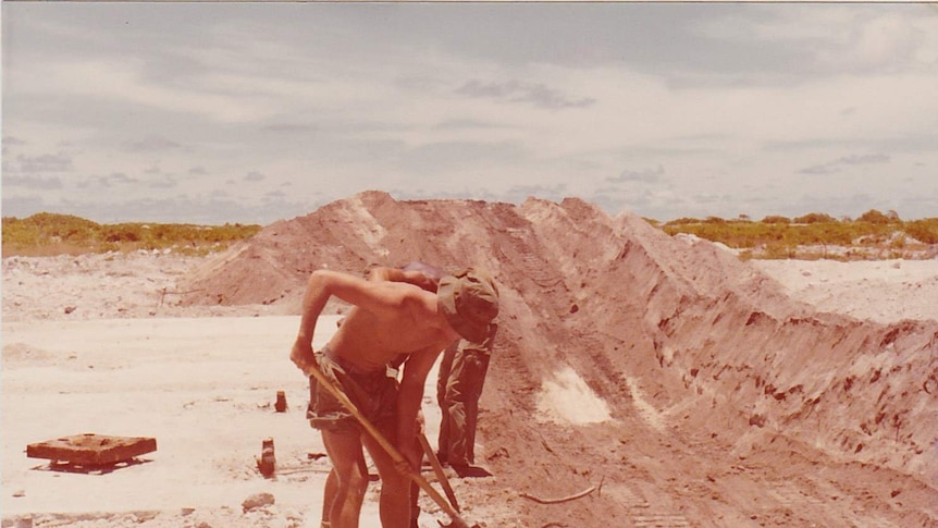 Shirtless soldiers dig up rubble with shovels next to a hole dug by machinery.