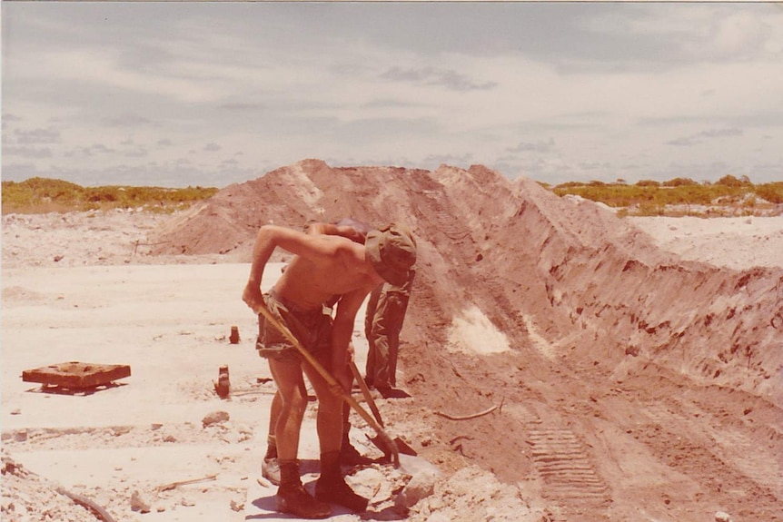 Shirtless soldiers dig up rubble with shovels next to a hole dug by machinery.