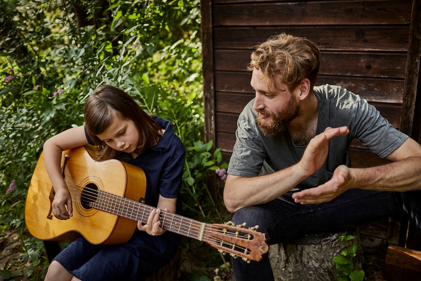 Father and daughter sit outside on a deck while the daughter plays guitar and he cheers her own. 