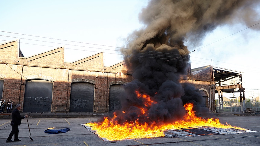 A man in black suit holds a shovel stands next to grid of artworks on fire in industrial area. Black smoke rises to the sky.