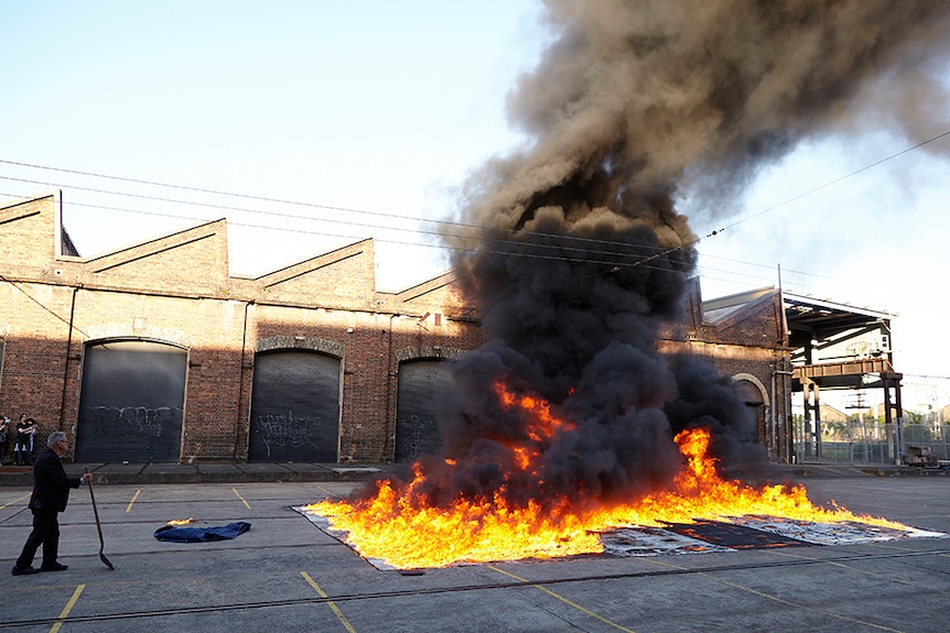 A man in black suit holds a shovel. He stands next to grid of artworks on fire in industrial area. Black smoke rises to the sky.