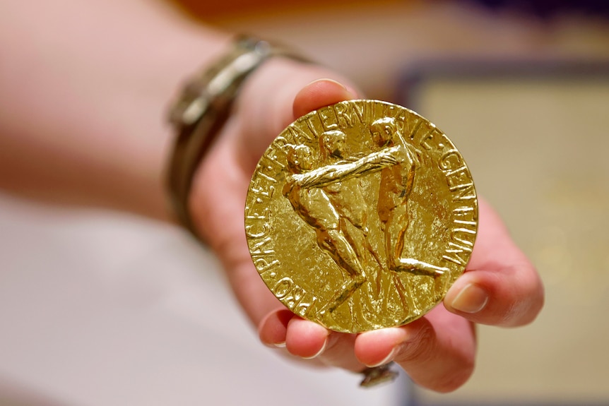 A white person's left hand holds a tennis-ball sized shiny gold medal before the camera