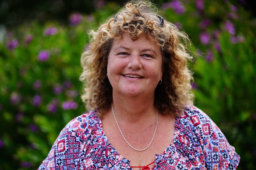 A woman smiling at the camera, standing in front of a bush with purple flowers