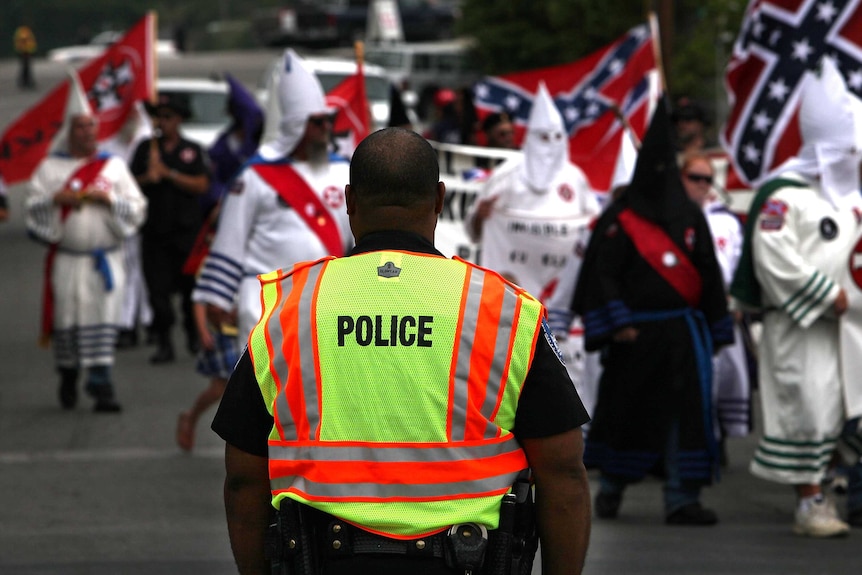 A black police officer watches as members of the Fraternal White Knights of the Ku Klux Klan participate in a rally.