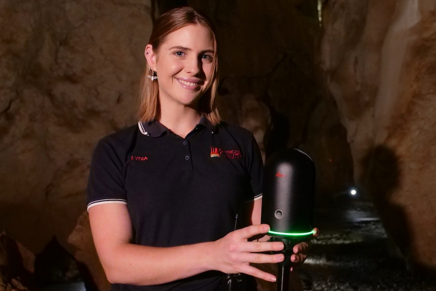 A young woman stands in front of a scan machine on a tripod in a cave, smiling