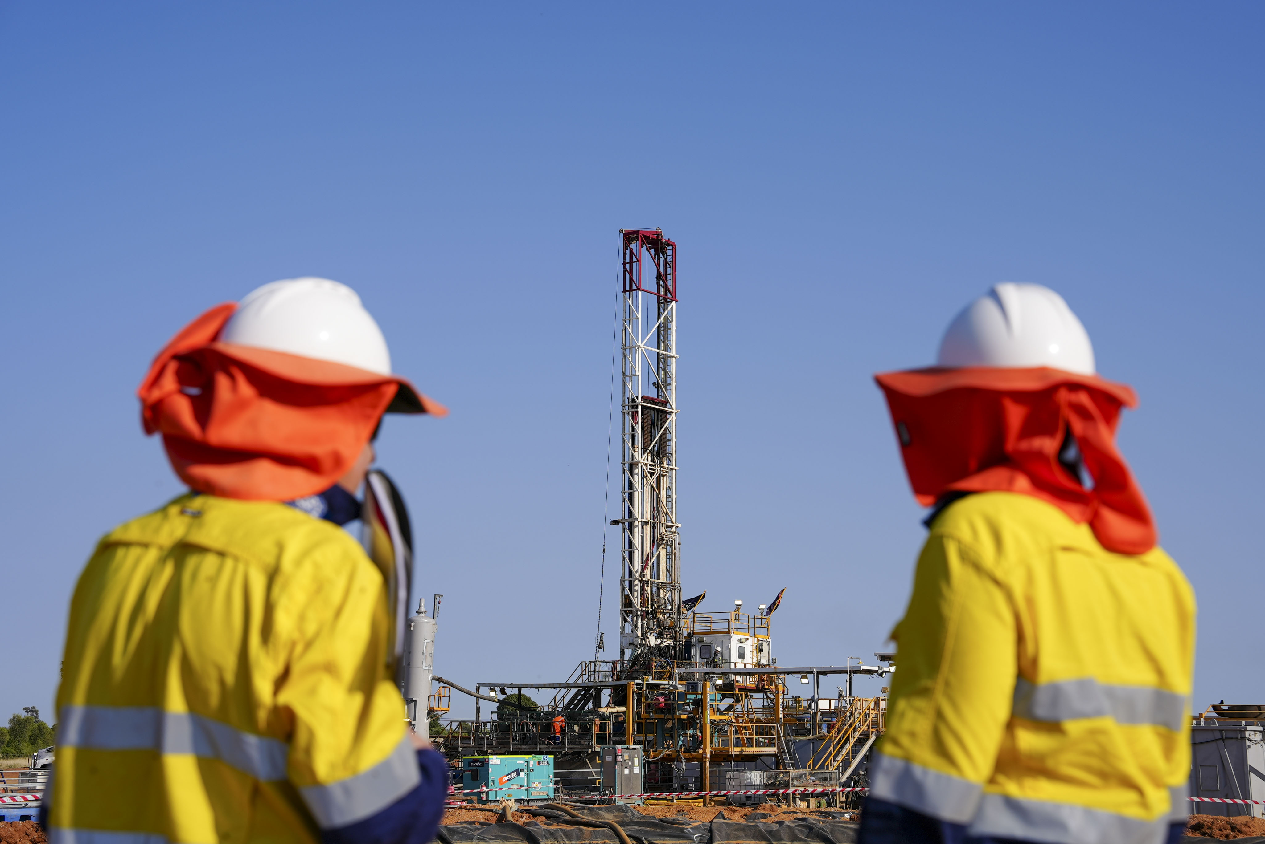 Workers in high-vis and hard hats stand in front of a gigantic drill rig that rises high into the sky.