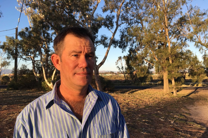 A white man in a blue striped shirt.  He has black hair and stands in a field with trees behind him.