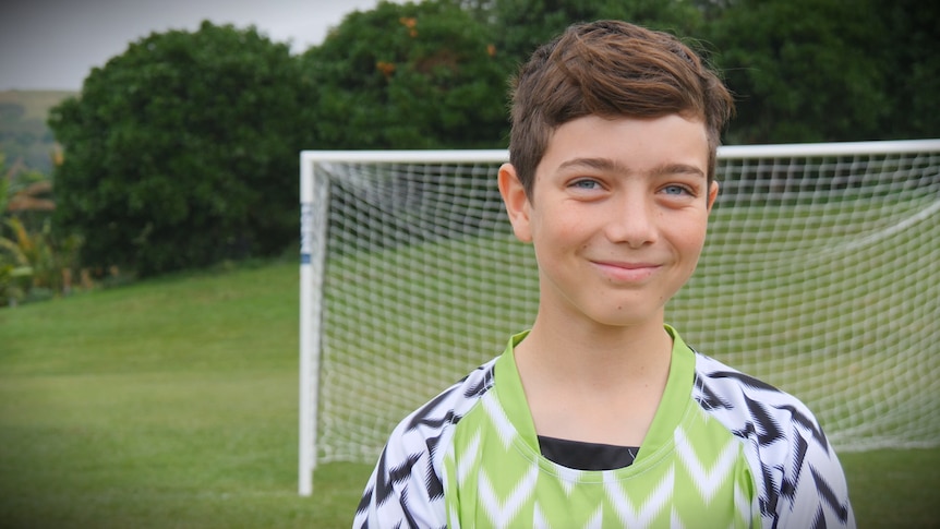 A 12-year-old boy in a green soccer jersey smiles with a soccer net in the background