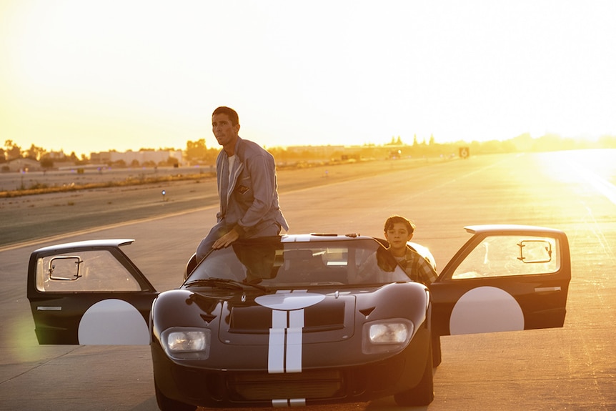 Christian Bale sits on roof of racing car while Noah Jupe peers from passenger seat, car sits on race track during sunset.