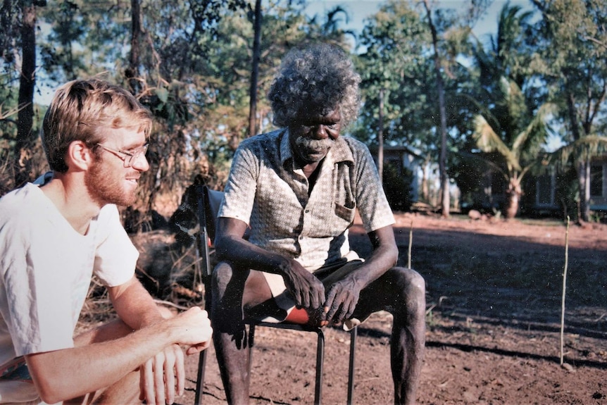 Two men sitting outside in the bush looking at the ground and into the distance on a sunny day.