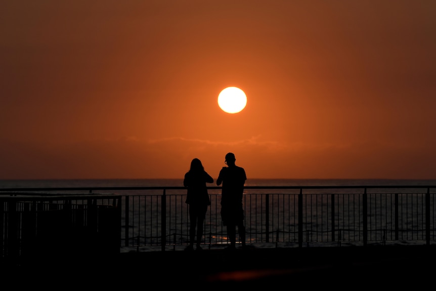 two people are seen up against a rail overlooking the water at a sydney beach on a hot day