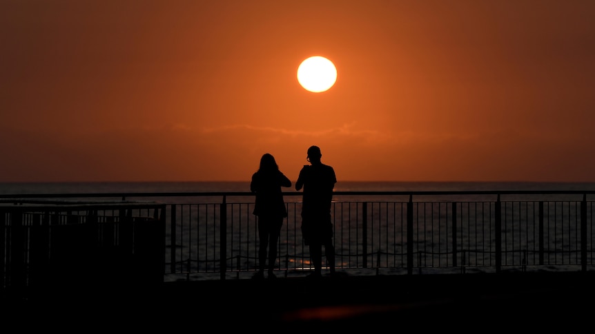 two people are seen up against a rail overlooking the water at a sydney beach on a hot day