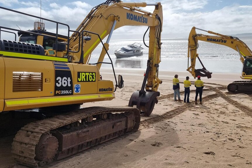 Two excavators on a beach in front of a sunken yacht
