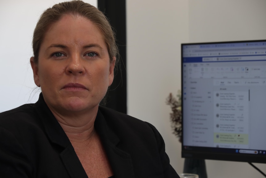 A photo of Hannah-Fincham Thomson in a black suit looking directly into the camera with a computer monitor in the background