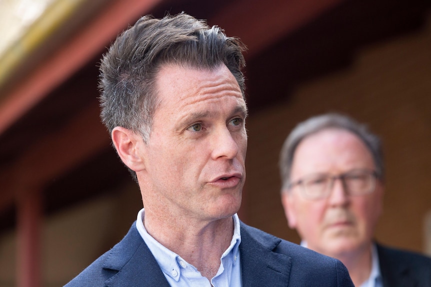 A Caucasian man with brown hair and a blue suit speaks to the media.