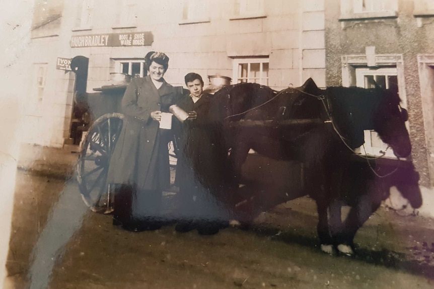 Two people stand in front of a horse and cart outside an old, sandstone shoe shop. The photo is in black and white.