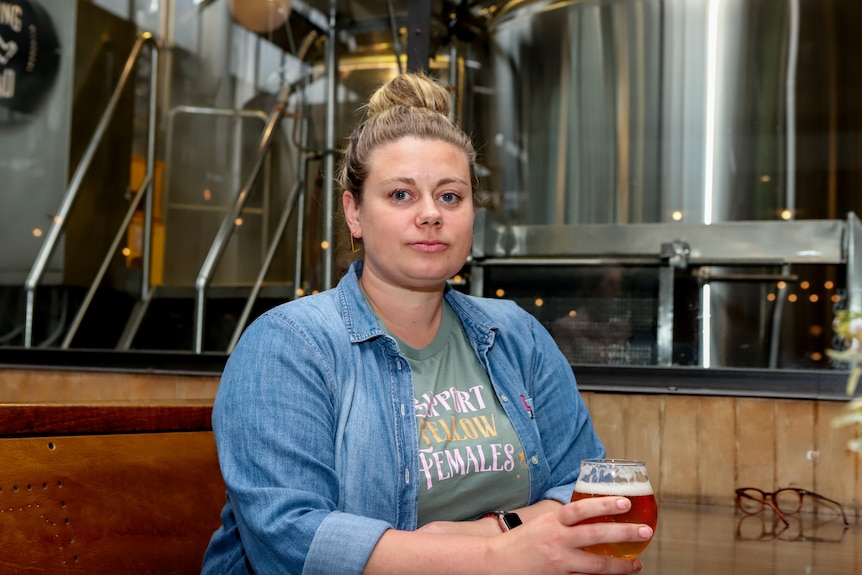 Woman with denim shirt and green t-shirt holds beer sitting at table with brewery in background 