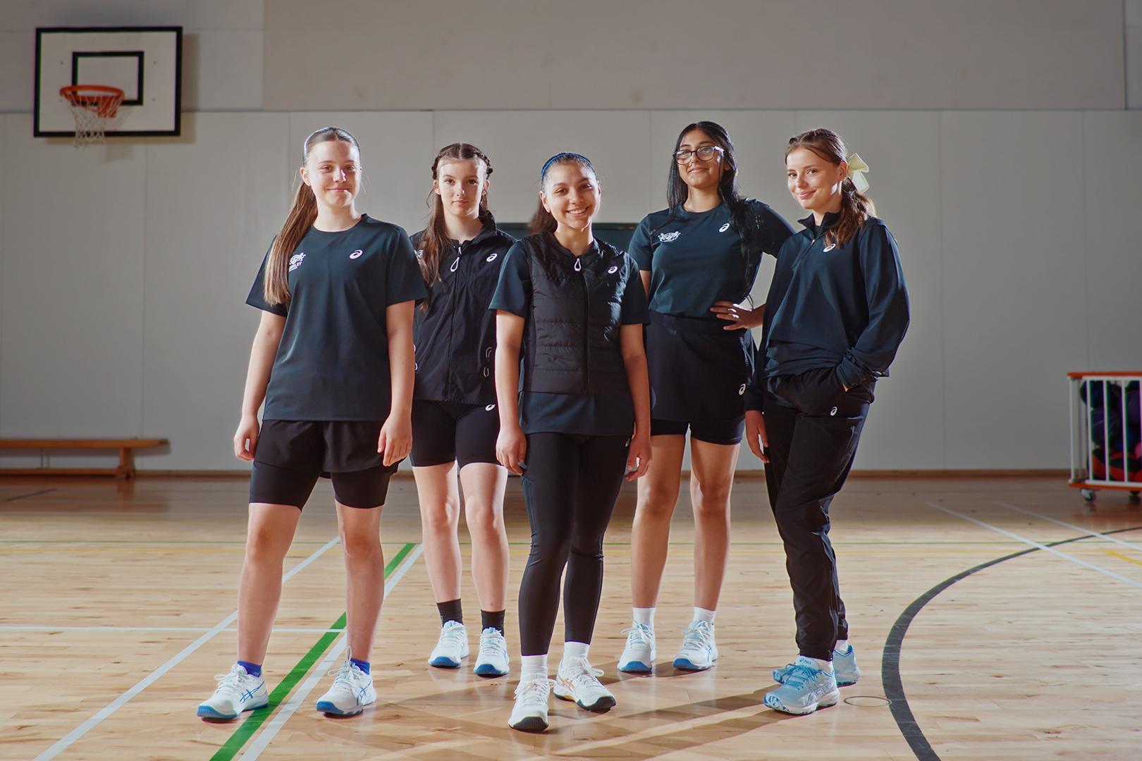 Grupo de mulheres vestindo roupas esportivas escuras fica na quadra de basquete.