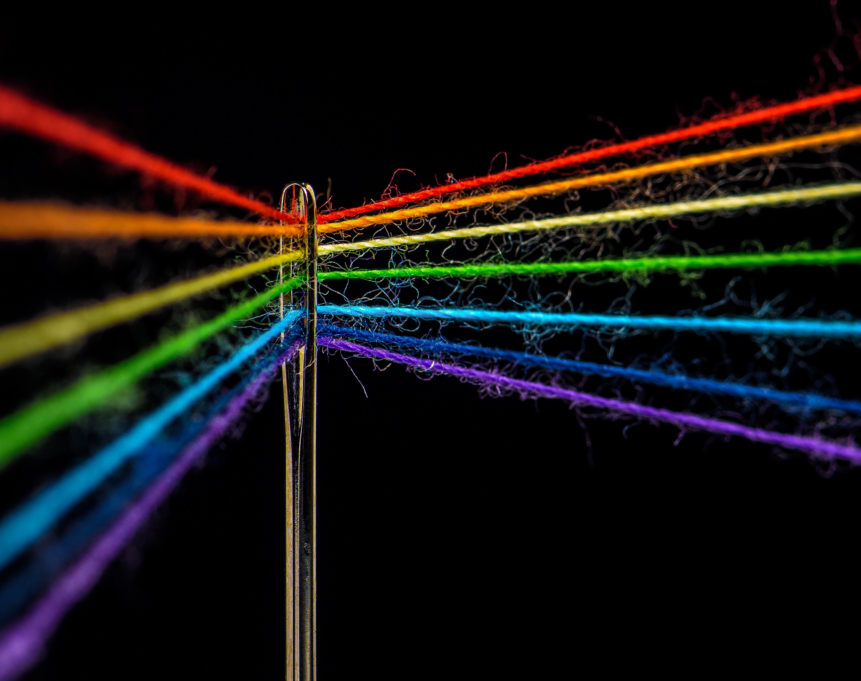 Macro image of multicoloured threads that form the colours of the rainbow. They are threaded through the eye of a sewing needle.